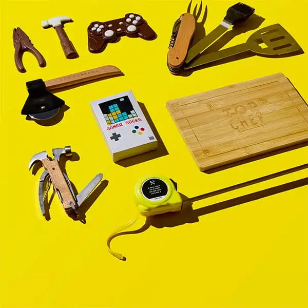 Flat lay on a yellow background featuring assorted Fathers Day gifts including tools, BBQ utensils, a wooden chopping board, novelty socks, and a tape measure